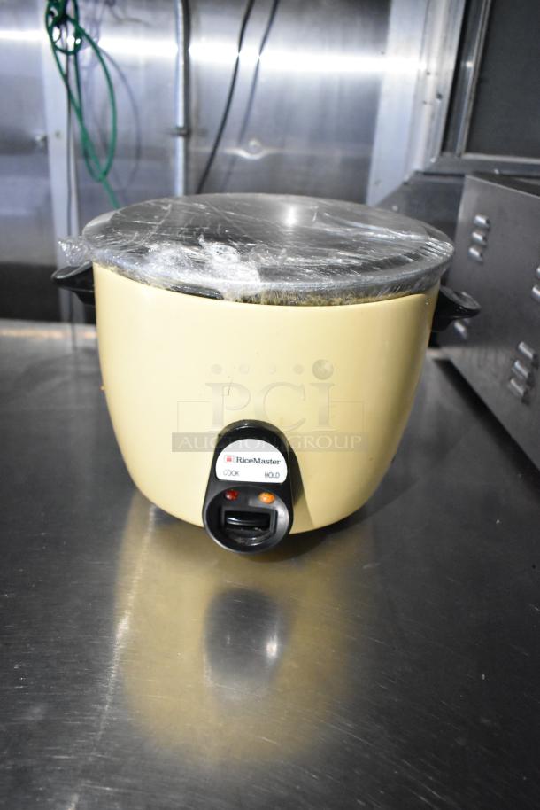 RiceMaster rice cooker with clear wrap on lid, placed on stainless steel surface. "Cook" and "Hold" functions visible.