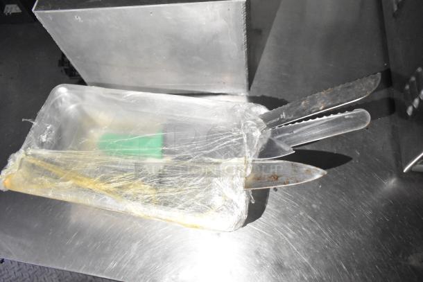 Plastic-wrapped metal tray with kitchen knives on a stainless steel surface.