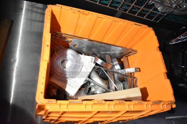 Orange storage bin containing metal parts, rolls of tubing, and packaging materials, indicating kitchen equipment components.