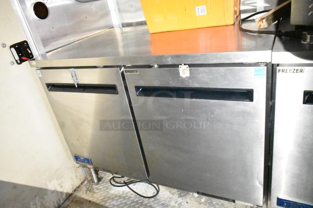 Mobile kitchen trailer interior with stainless steel Arctic Air prep tables, refrigerator, freezer, and diamond plate flooring.