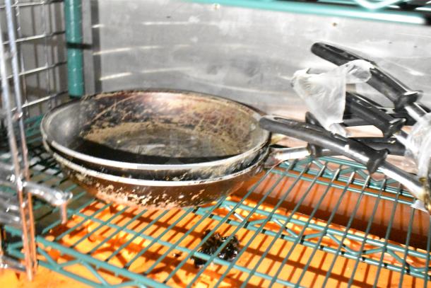 Stack of worn frying pans with plastic-wrapped handles on a green wire shelf, part of mobile kitchen inventory.
