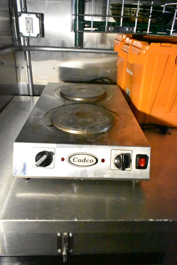 Cadco double burner hotplate on stainless steel counter inside mobile kitchen trailer with orange storage bins.