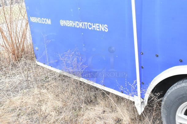 Blue mobile kitchen trailer with visible markings: "NBRHD.COM" and "@NBRHDKITCHENS." Fiberglass body near dry grass.
