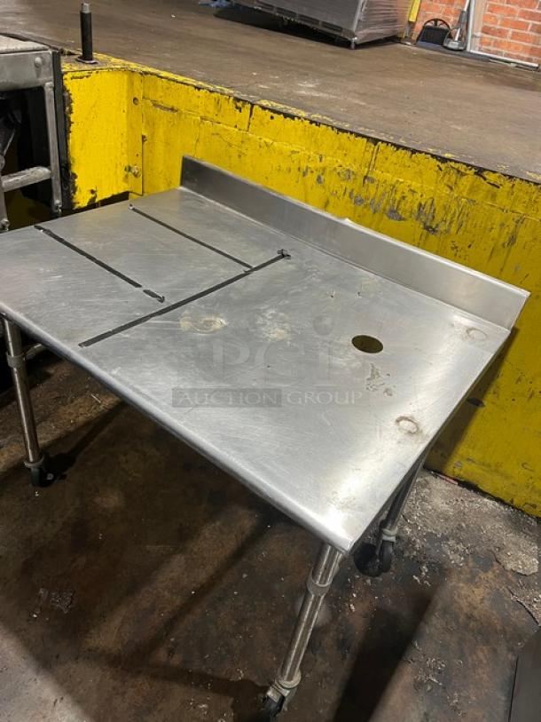 Commercial stainless steel worktop table with upturned backsplash, mounted on casters. Features include hole cutouts and rails; used condition.