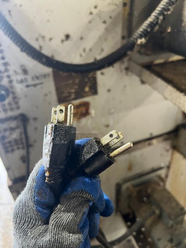 A gloved hand holds two power plugs against a background showing part of a commercial kitchen appliance.