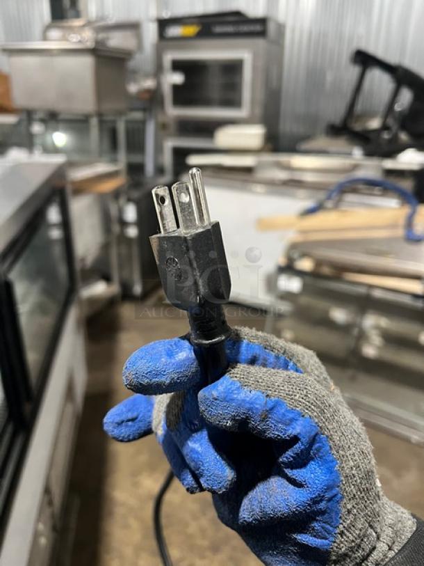 Close-up of an electrical plug held by a gloved hand, in a commercial kitchen setting.