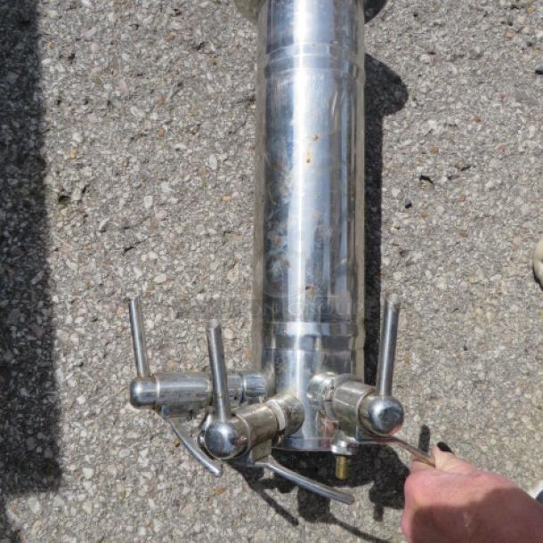 Stainless steel wine tower with three taps, polished finish, outdoor setting, shows minor wear, ideal for home bars.