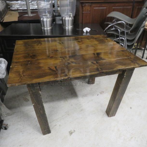 A solid wooden table with a rustic finish and four sturdy legs, exhibiting a natural wood grain. Appears in used condition.