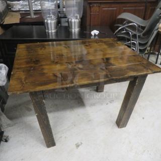 A solid wooden table with a rustic finish and four sturdy legs, exhibiting a natural wood grain. Appears in used condition.