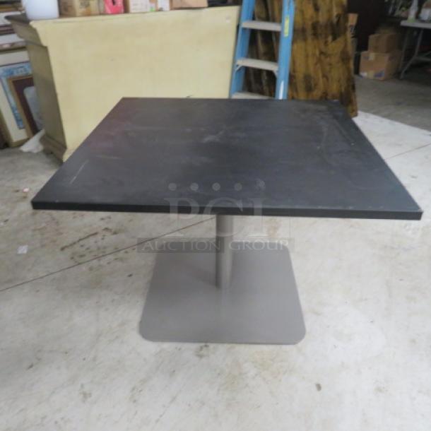 Square black laminate table top with pedestal base, 42x42x30 inches, shows light wear, sturdy metal base.