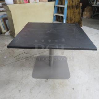 Square black laminate table top with pedestal base, 42x42x30 inches, shows light wear, sturdy metal base.