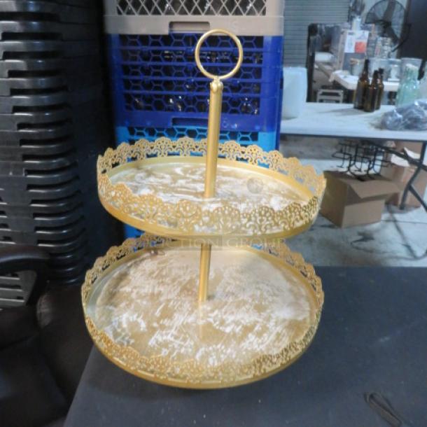 Two-tier gold display stand with ornate edging, loop handle, and metallic finish. Slight wear visible on tiers.