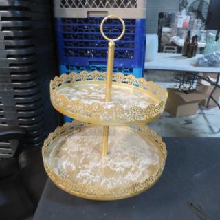Two-tier gold display stand with ornate edging, loop handle, and metallic finish. Slight wear visible on tiers.