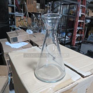 Clear glass carafe with wide base, narrow neck, excellent condition, Item 6XBID, displayed in warehouse setting.