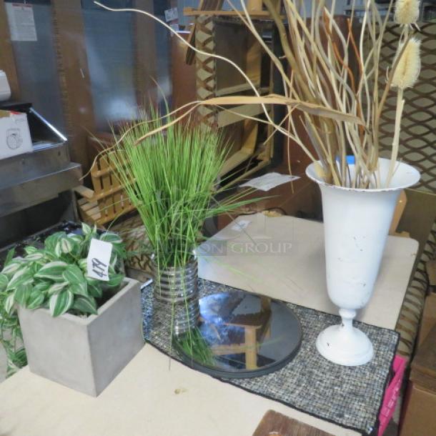 Assorted decor lot featuring faux plants in vases, including metal urn and concrete pot, displayed on patterned cloth.