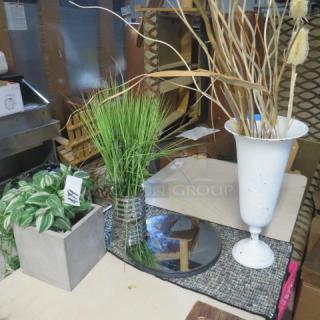Assorted decor lot featuring faux plants in vases, including metal urn and concrete pot, displayed on patterned cloth.
