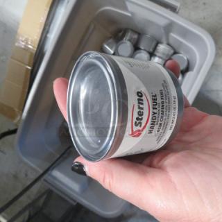 Hand holding a can of Sterno Handy Fuel. Features visible label with brand name and description. Other cans in a plastic bin.