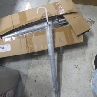 A case of new clear umbrellas in an open cardboard box, each with white handles, pristine condition, ready for use.