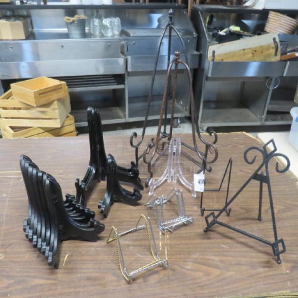 A variety of easels, including metal, wooden, and clear plastic stands, shown on a table, all in used condition.