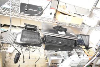 Alt Text: 
Auction items on a wire shelf, including two black cash drawers and five POS monitors. Tags visible with item numbers.