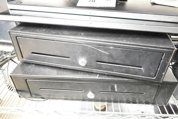 Two black cash drawers with key locks, slight wear. Stacked on shelving, part of auction lot including 5 POS monitors.