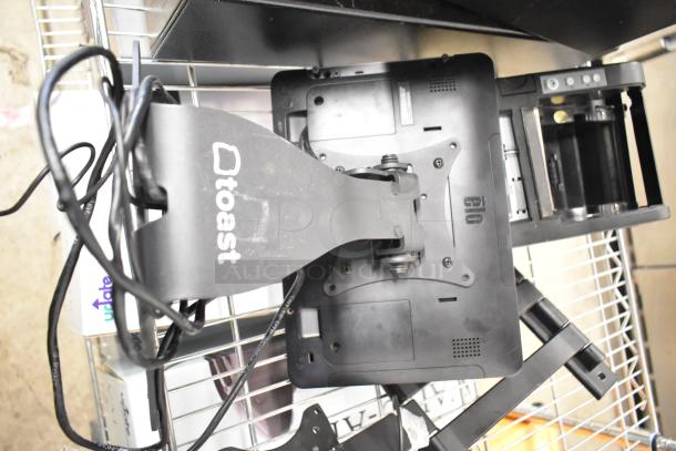10 various items including Toast POS monitor, cash drawer, cables, and components on wire rack. Good condition.