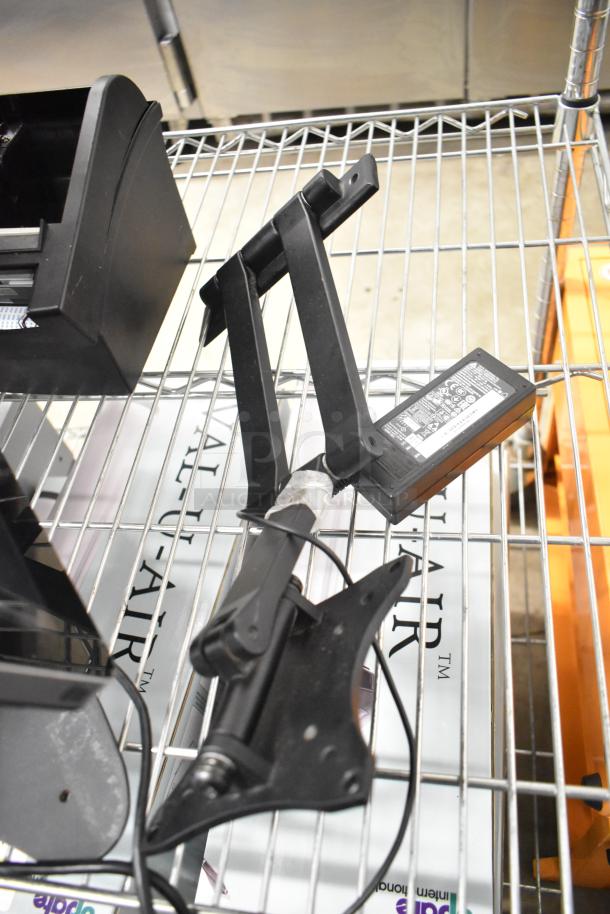 Assorted auction items including a black cash drawer and monitor stand, in used condition, on a metal rack.