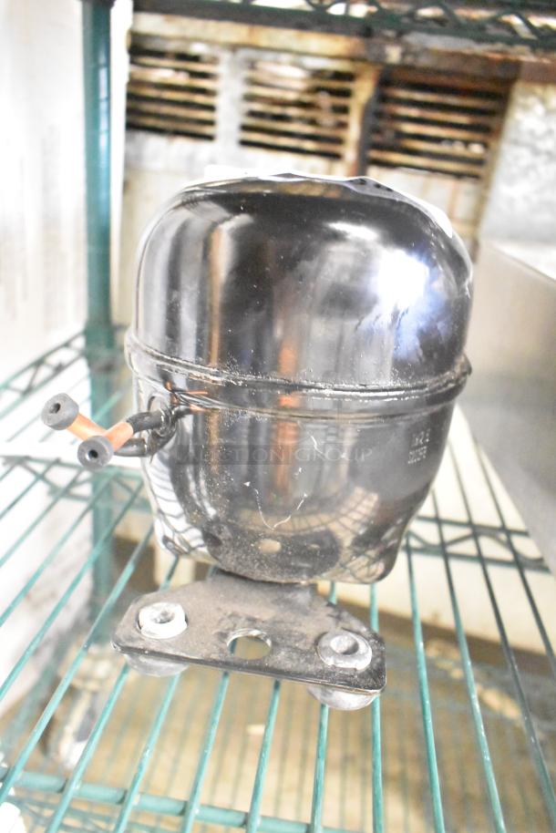 Cubigel NUT55NR Compressor, 151-127V, 1 Phase. Black casing, mounted on a green wire shelf. Some visible surface wear.