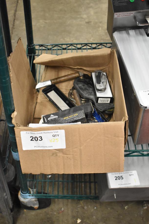 Cardboard box containing various items including knives, brass knuckles, and handcuffs. Condition appears used. Tag reads "203 QTY V25."