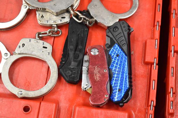 Various items including silver handcuffs, black and red pocket knives, and brass knuckles on a red surface. Knives feature unique patterns.
