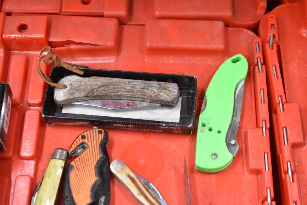 Various pocket knives on a red tray; includes wood-handled and green plastic-handled knives.