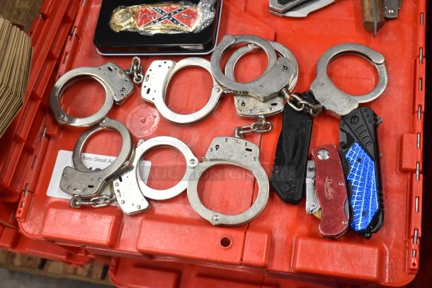 A collection of handcuffs, brass knuckles, and pocket knives on a red crate, showing wear and diverse designs.