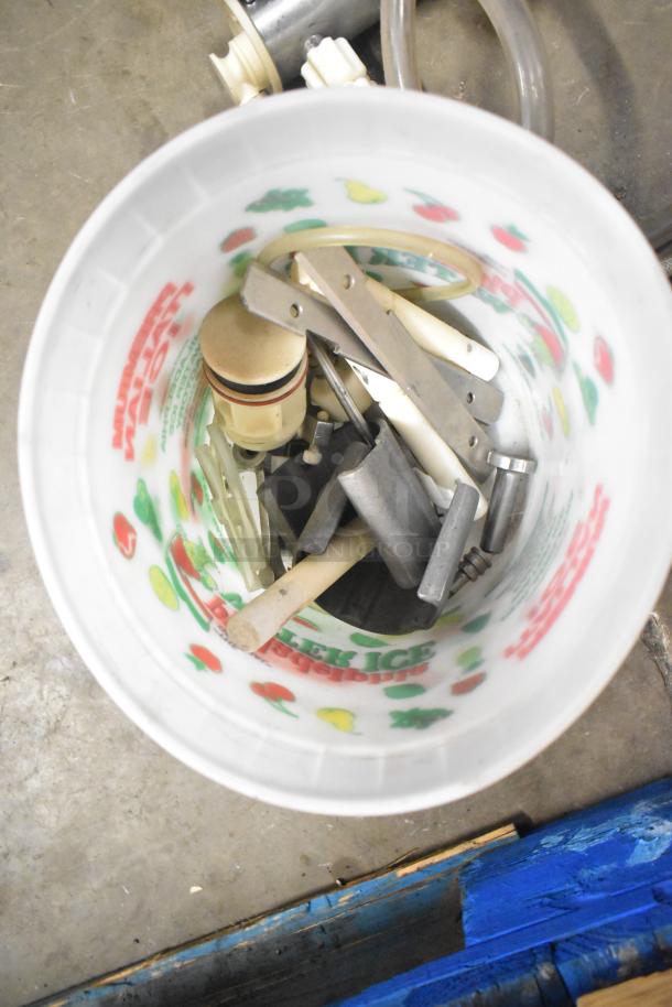Tiered lot of various machine parts in a bucket. Includes Turbochef kickplate, Taylor ice cream machine drip tray, carbonated beverage machine drip tray. Items unassembled, showing moderate wear. Bucket with colorful fruit design.