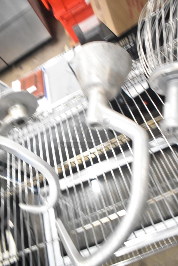 15 attachments for Hobart Mixer, including dough hooks, paddle whips, whisks. Various metal pieces on wire rack.