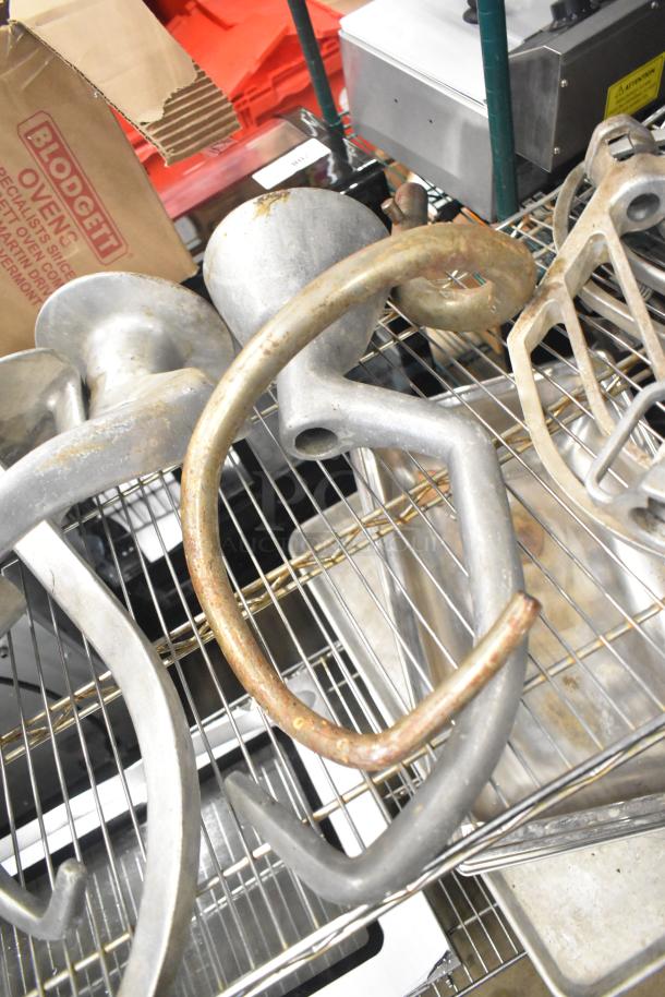 15 Hobart mixer attachments, including dough hooks, paddle whips, and whisk, in used condition on wire shelf.
