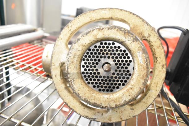 Vintage metal meat grinder attachment with rust and wear, featuring circular grinding plate and bolt, positioned on grill rack.