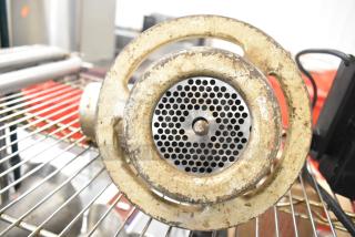 Vintage metal meat grinder attachment with rust and wear, featuring circular grinding plate and bolt, positioned on grill rack.