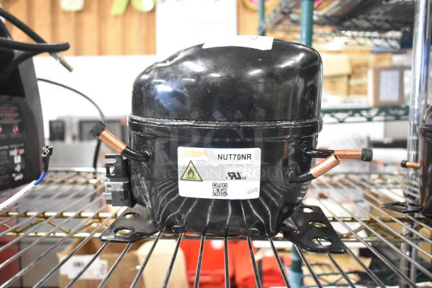 Cubigel NUT70NR compressor, 115-127V, 1 phase, black, thermally protected, mounted on metal shelf, product label visible.