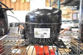 Cubigel NUT70NR compressor, 115-127V, 1 phase, black, thermally protected, mounted on metal shelf, product label visible.