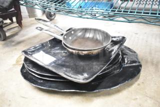 Stack of black baking pans and two stainless steel skillets, used condition, on a concrete floor. White tag visible.