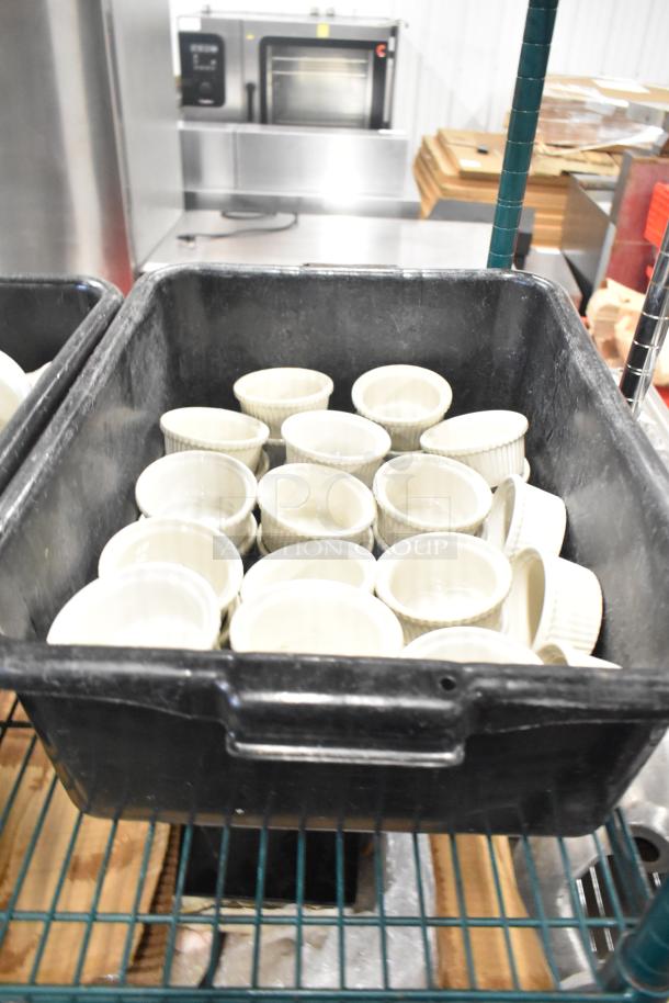 Black poly bus bin containing multiple white ceramic ramekins on a metal shelving unit. Auction tier lot.