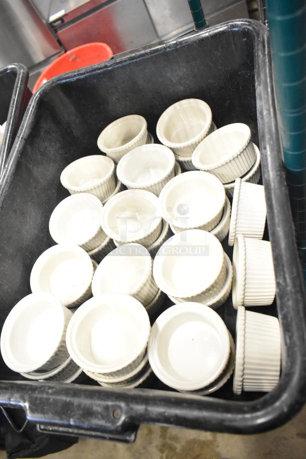 Black poly bus bin filled with multiple white ceramic ramekins. Ramekins are ribbed, round, and appear in good condition.