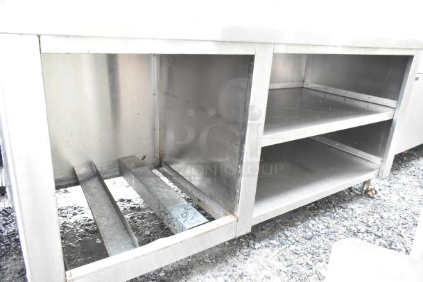 Commercial stainless steel table with overhead and under shelves, visible wear, suitable for kitchen or workshop use.