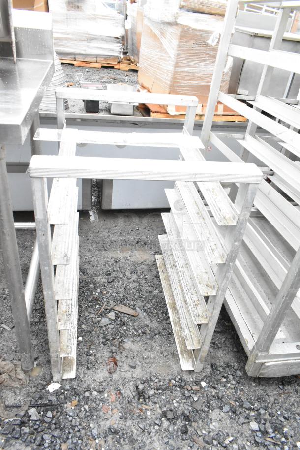 Commercial pan rack with multiple shelves, aluminum construction, outdoor setting, shows signs of wear and use.