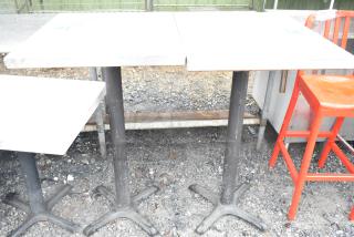 Two bar-height tables with white tops on black metal bases, outdoor setting, includes red bar stool, moderate wear.
