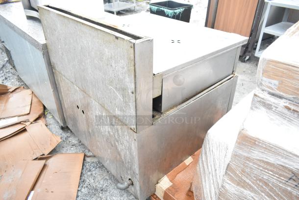 Commercial floor-style braising pan, stainless steel, visible wear and weathering, situated outdoors, no visible labels.