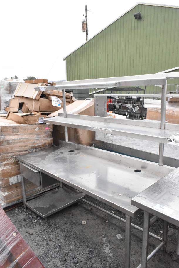 Commercial table with two-tier overshelf and drawer. Stainless steel, sturdy structure. Some surface wear visible.