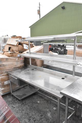 Commercial table with two-tier overshelf and drawer. Stainless steel, sturdy structure. Some surface wear visible.