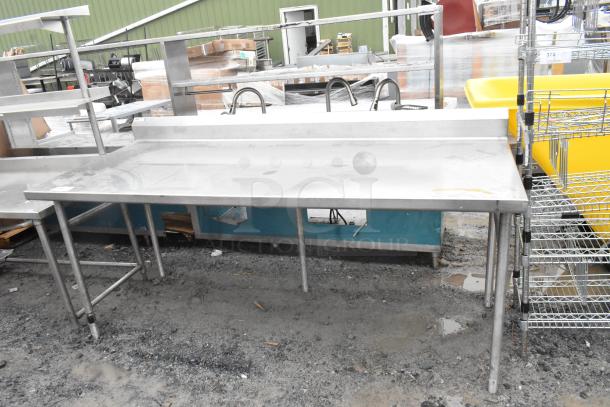 Stainless steel commercial table with multiple legs, integrated backsplash, and sturdy construction. Signs of use visible.