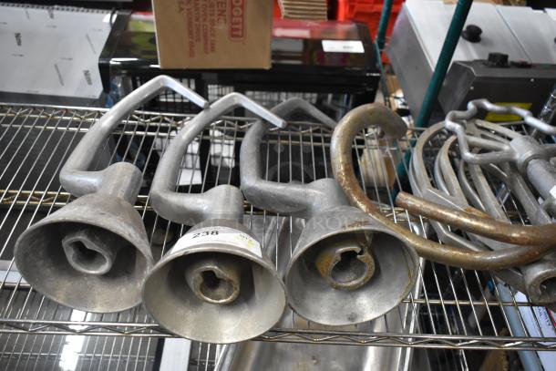 Four various dough hook attachments for Hobart mixers on a metal shelf, in used condition, with visible wear.
